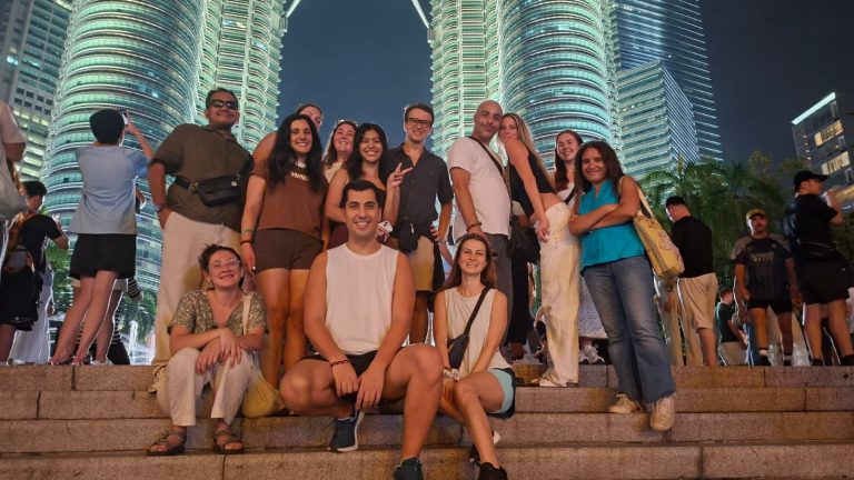 A Travelogue Guesthouse group with their arms up, the Petronas Twin Towers behind them