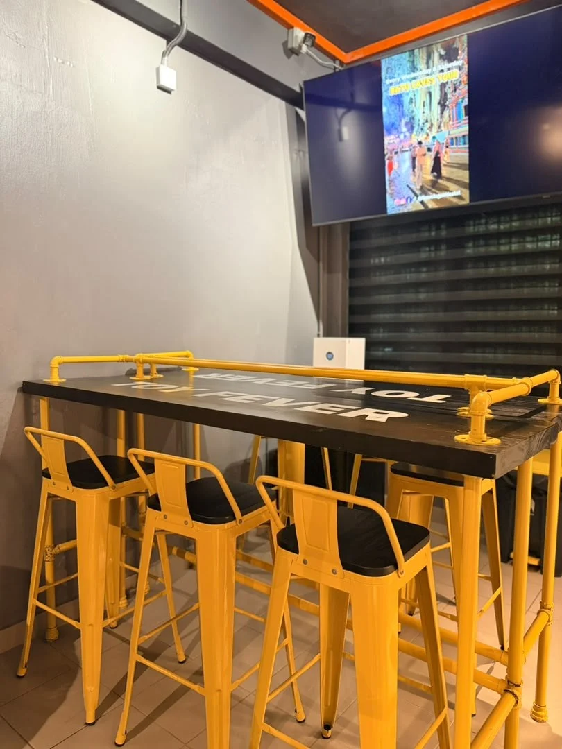 A black bar table with yellow stools under a wall-mounted screen in the common area at Travelogue Guesthouse