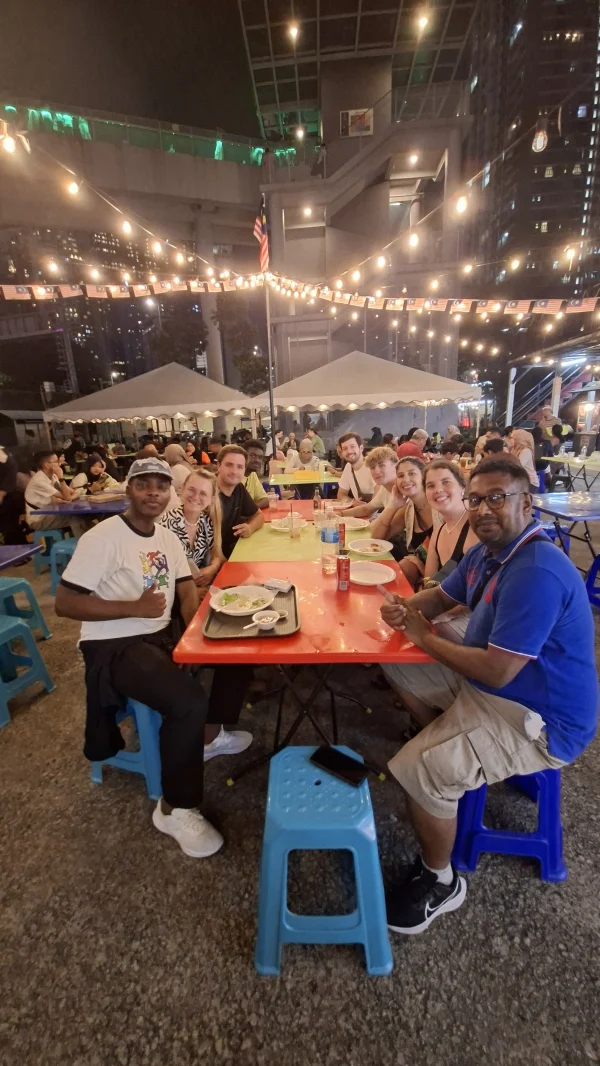 Travelogue Guesthouse guests around a long table of street food under string lights at night, Kuala Lumpur