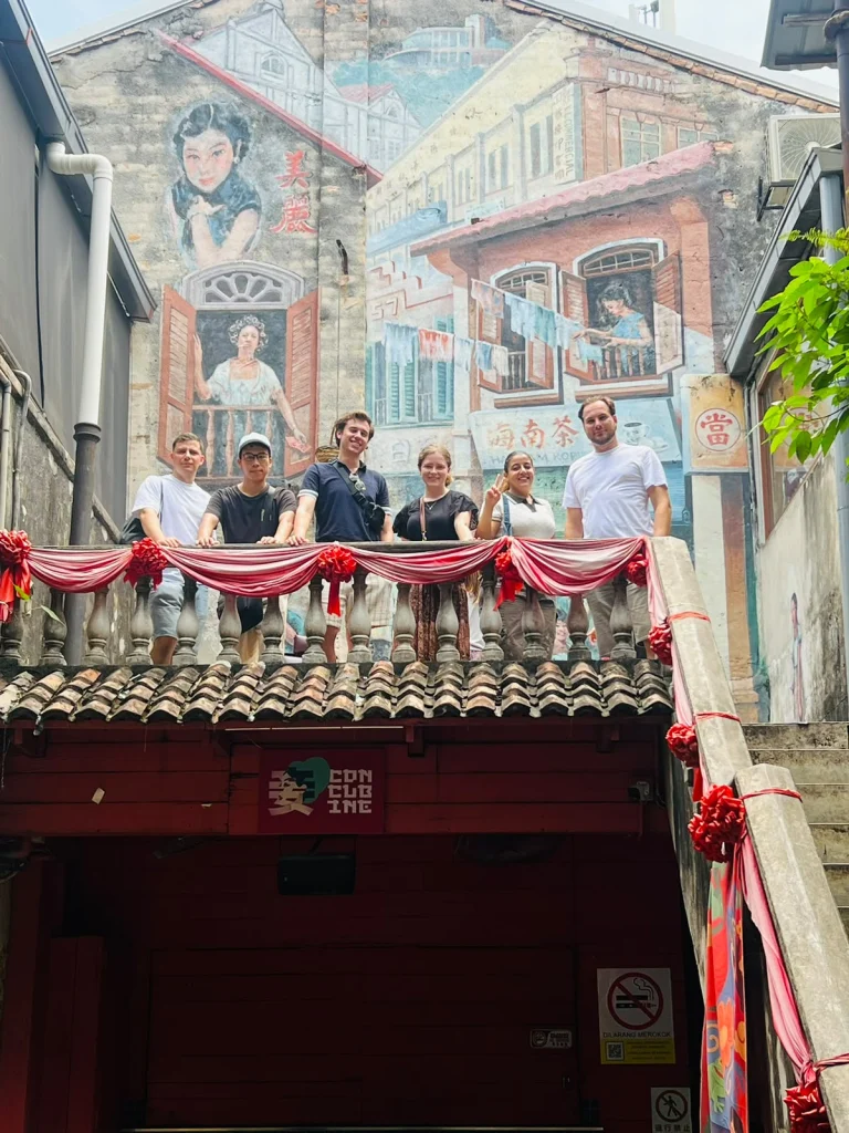 Travelogue Guesthouse guests on the balcony at Kwai Chai Hong, red fabric swagged along the balustrade and the lane's shophouse murals on the wall behind them