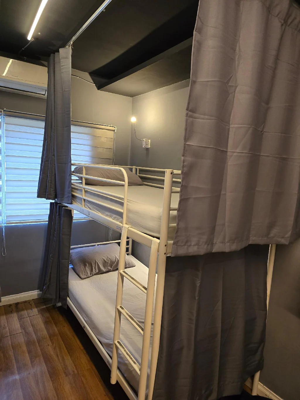Two bunks in the male dormitory at Travelogue Guesthouse, each with a grey privacy curtain, a reading light and a socket, beside a shuttered window