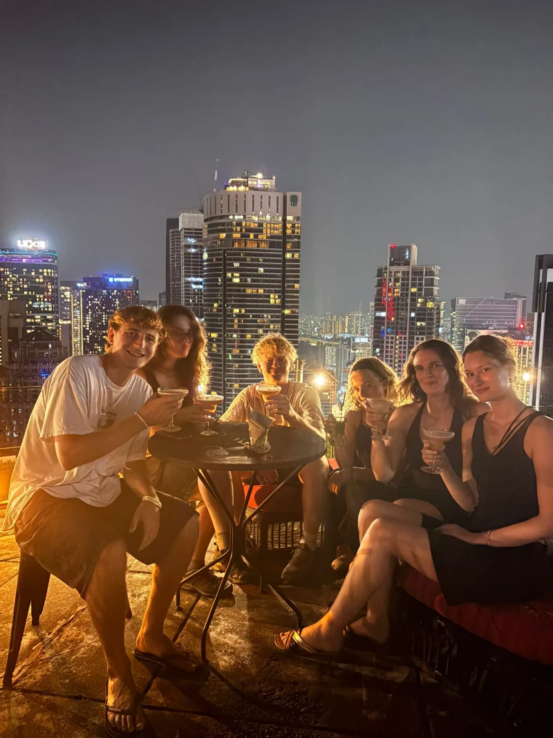 Travelogue Guesthouse guests with drinks at a rooftop bar table above the lit Kuala Lumpur skyline at night