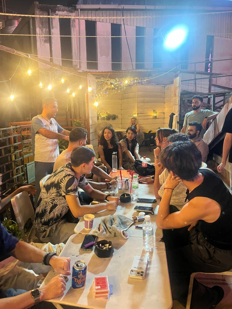Guests sitting along a long table on the Travelogue Guesthouse roof under the string lights, drinks and cards on the table