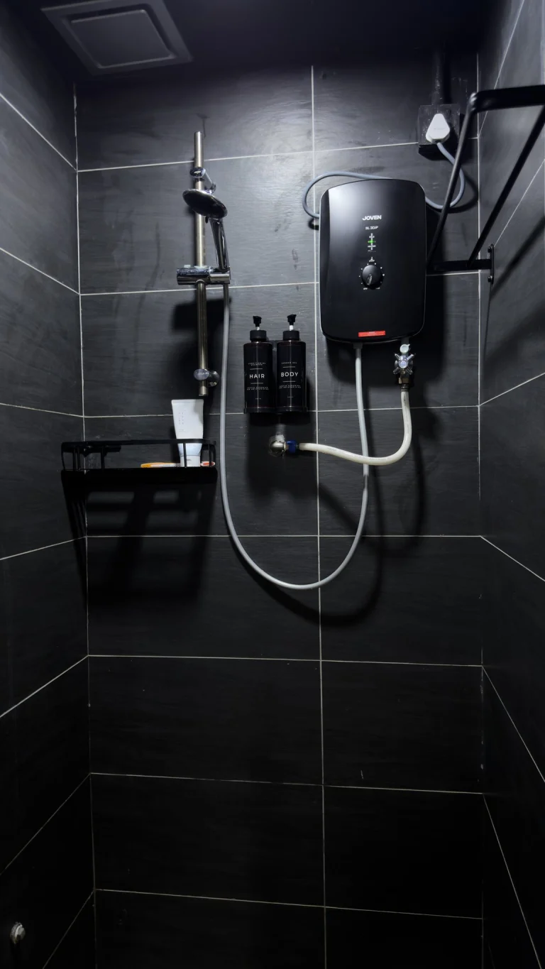 A shared shower cubicle at Travelogue Guesthouse, dark tiled, with an instant water heater, a hand shower on a rail and shampoo and body wash on a shelf
