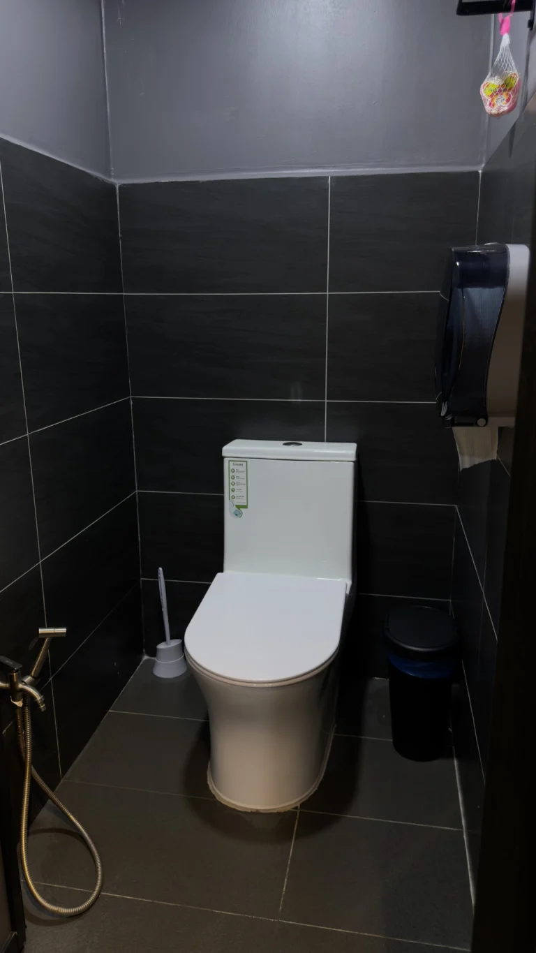 A shared toilet cubicle at Travelogue Guesthouse, dark tiled, with a bidet spray beside the toilet
