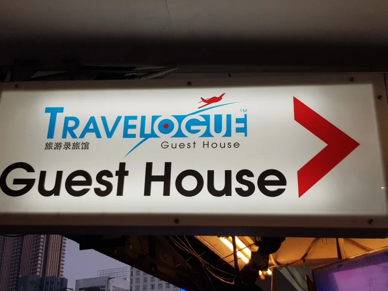 The lit Travelogue Guest House sign above the pavement on Jalan Bukit Bintang, with a red arrow on it pointing towards the entrance