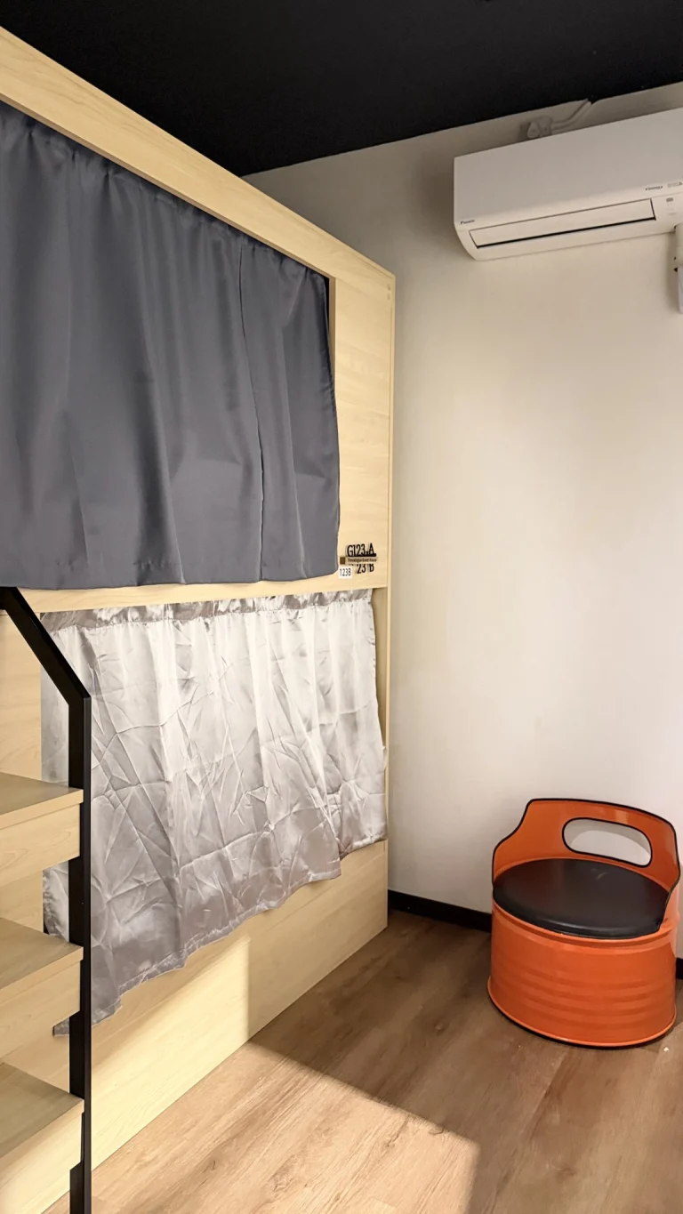 A pod bunk with a grey curtain above and a pale one below, beside an orange stool, in the female dormitory at Travelogue Guesthouse